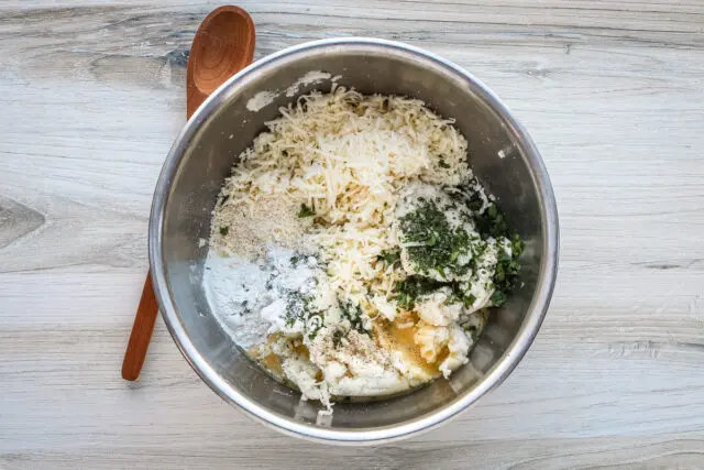 Combining the mashed potatoes with the remaining croquette ingredients, including cheese, herbs, flour, an egg, and seasonings.