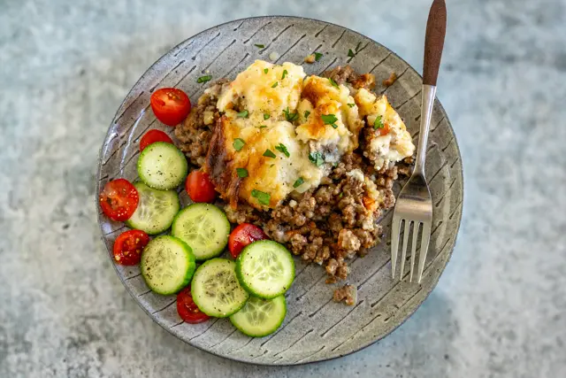 A serving of beef and potato casserole on a plate with cucumbers and a parsley garnish