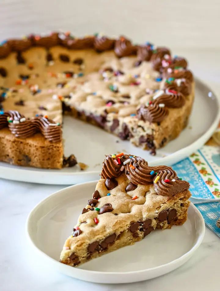 A slice of chocolate chip cookie cake with piped chocolate frosting and rainbow sprinkles.