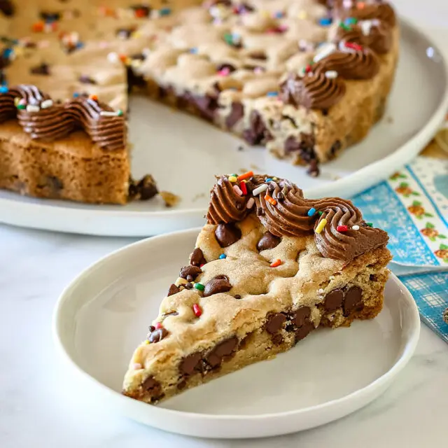 A slice of chocolate chip cookie cake with piped chocolate frosting and rainbow sprinkles.