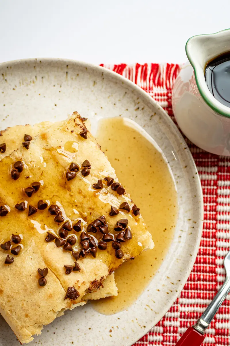 Slices of sheet pan pancakes on a plate with chocolate chip topping and syrup on the side.