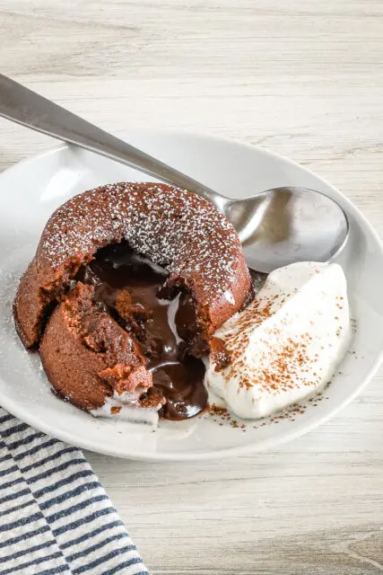 A chocolate fondant, also known as molten lava cake, on a plate with freshly whipped cream and a light dusting of cocoa and powdered sugar.