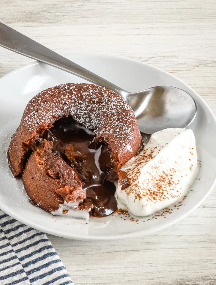 A chocolate fondant, also known as molten lava cake, on a plate with freshly whipped cream and a light dusting of cocoa and powdered sugar.