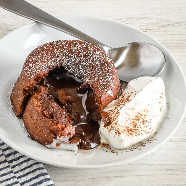 A chocolate fondant, also known as molten lava cake, on a plate with freshly whipped cream and a light dusting of cocoa and powdered sugar.