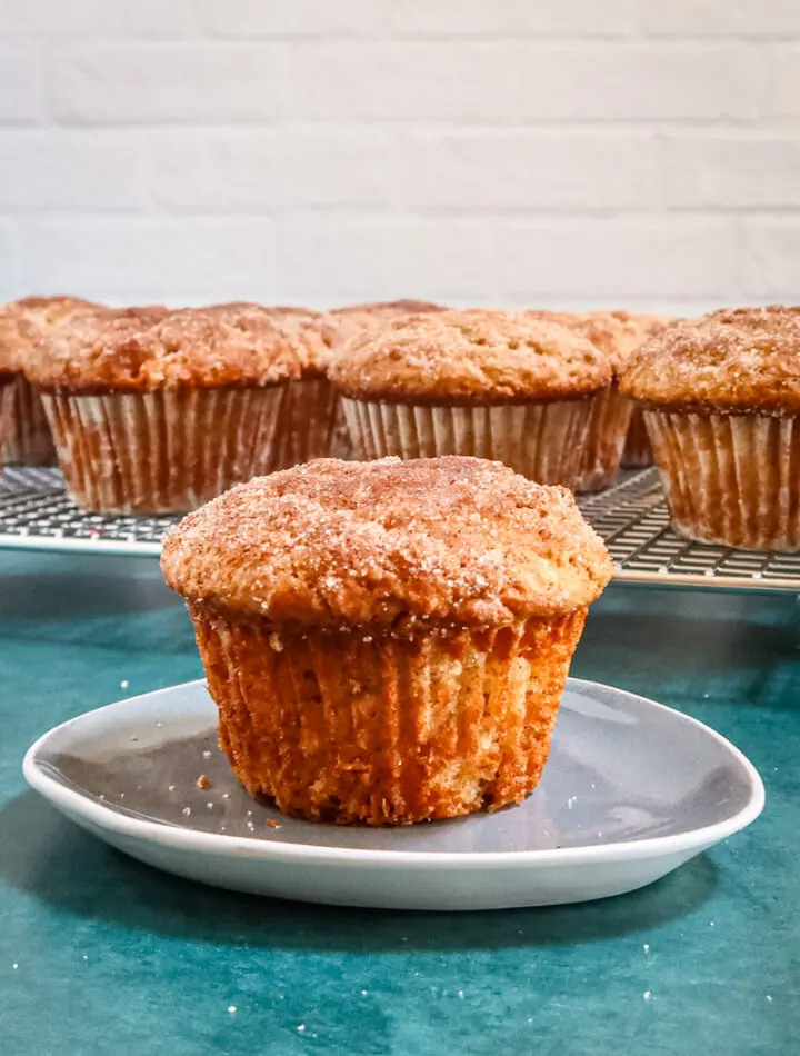 A cinnamon muffin on a small plate with the rack of muffins behind it.