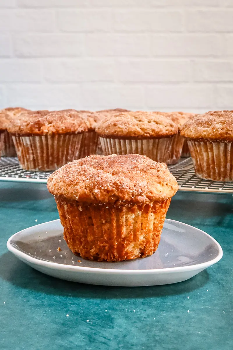 A cinnamon muffin on a small plate with the rack of muffins behind it.