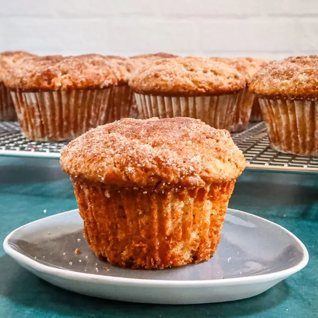 A cinnamon muffin on a small plate with the rack of muffins behind it.