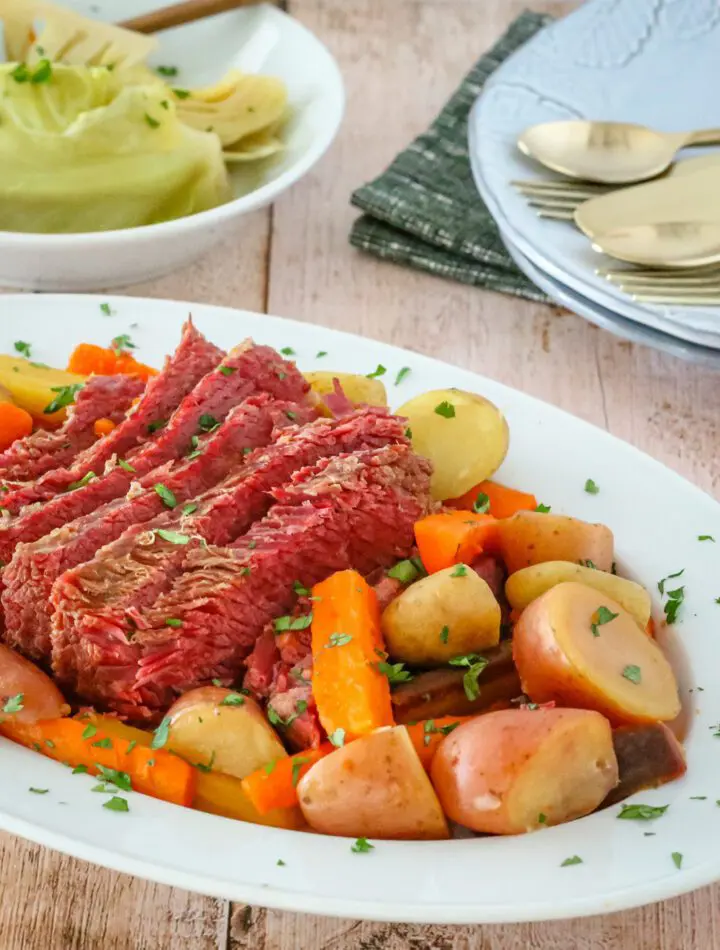 corned beef on a platter with potatoes, carrots, and cabbage on a plate in the background