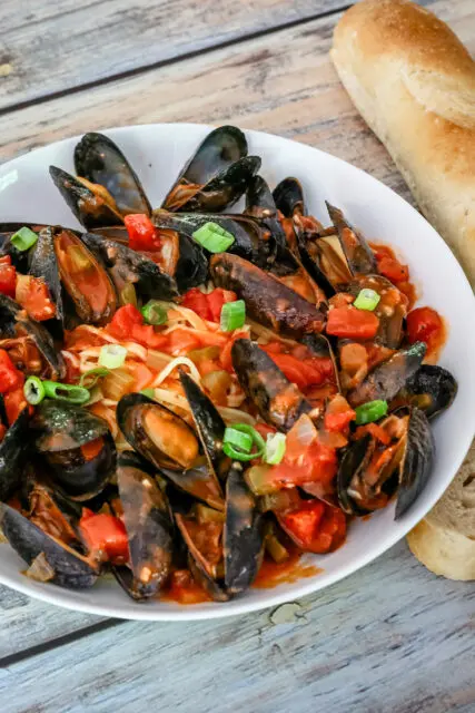 A bowl of mussels with Creole sauce, linguine, and a baguette on the side.