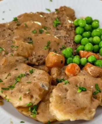 cube steak with mushrooms and gravy shown on a plate with potatoes, peas, and carrots