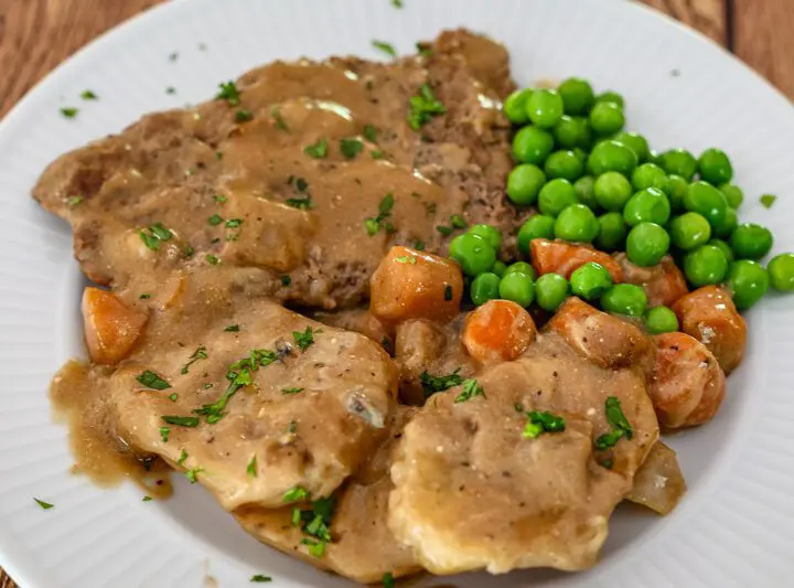 cube steak with mushrooms and gravy shown on a plate with potatoes, peas, and carrots