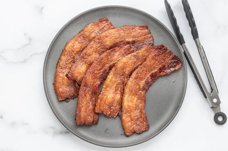 A plate with baked bacon and tongs on the side.