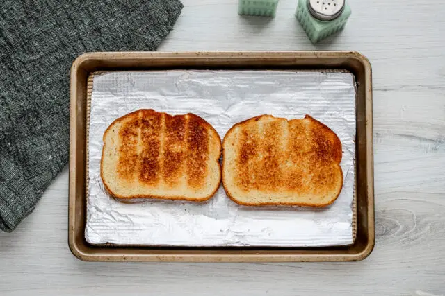 Two slices of toasted, lightly buttered bread are arranged on a foil-lined baking sheet.