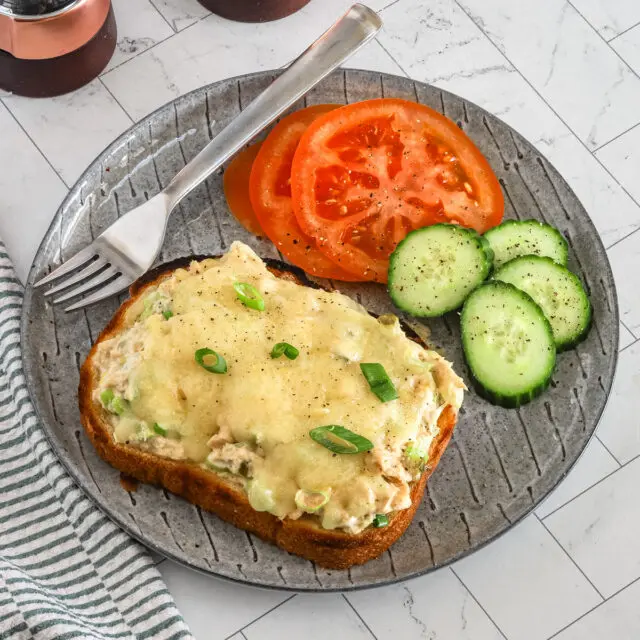 An elevated tuna melt on a plate with some sliced tomatoes, cucumbers, and a green onion garnish.