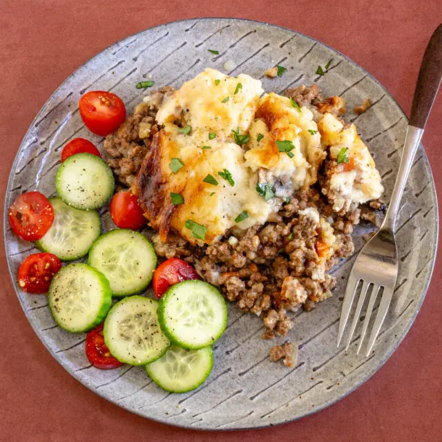 Ground beef casserole with mashed potato topping on a plate with some cucumbers and tomatoes.