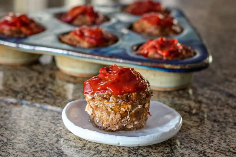 fun meatloaf muffins with 1 muffin on a plate with ketchup topping and more in a muffin baking dish in the background.
