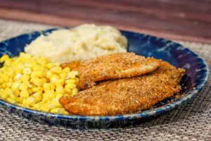 Oven fried chicken breasts on a plate with corn and mashed potatoes.