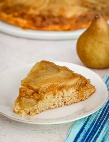 A slice of pear upside-down cake on a plate with a pear in the background.