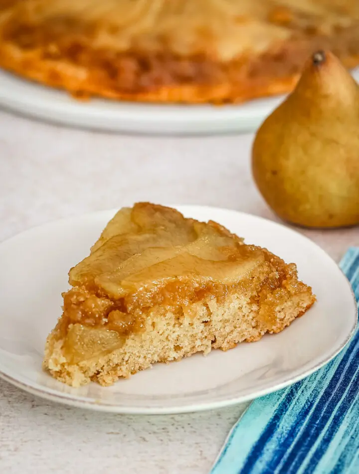 A slice of pear upside-down cake on a plate with a pear in the background.