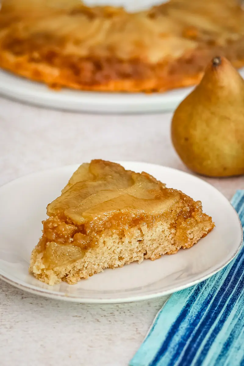 A slice of pear upside-down cake on a plate with a pear in the background.