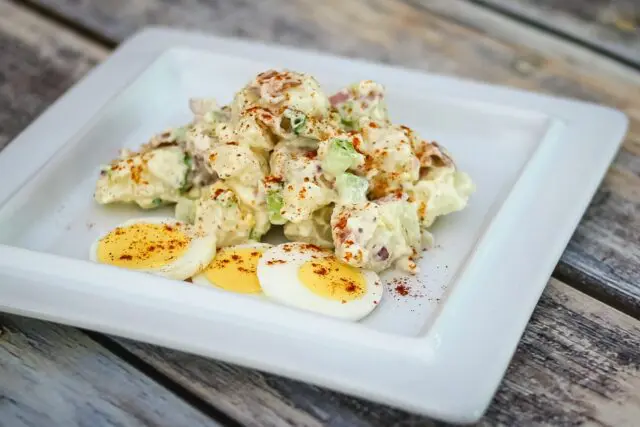 A bacon and egg potato salad on a serving plate with some sliced hard-boiled eggs on the side.