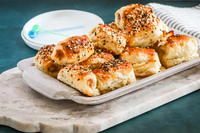 A serving tray with baked puff pastry pigs in a blanket with small appetizer plates and picks for serving.