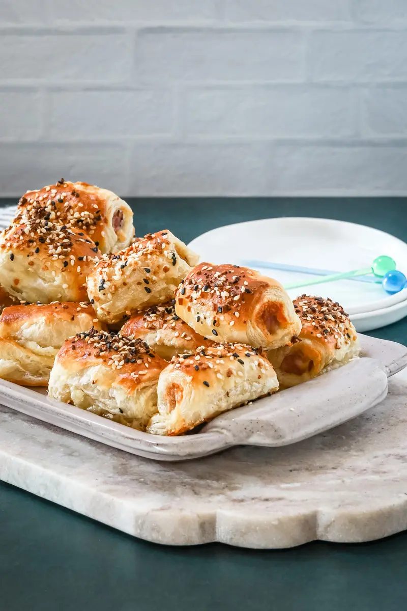 A serving tray with baked puff pastry pigs in a blanket with small appetizer plates and picks for serving.