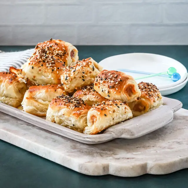 A serving tray with baked puff pastry pigs in a blanket with small appetizer plates and picks for serving.