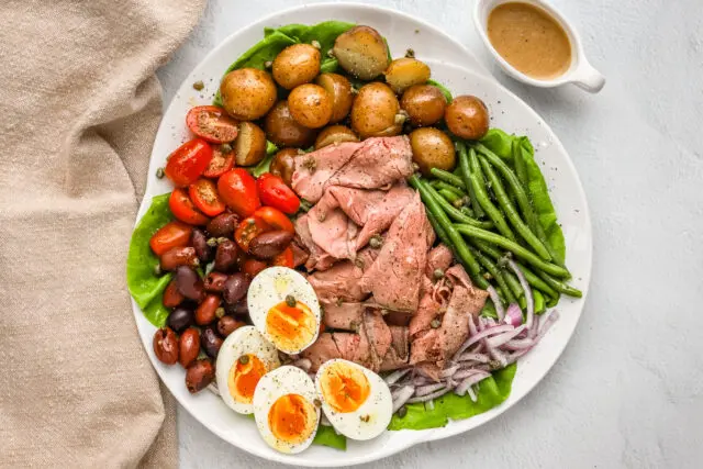 Beef nicoise ingredients are assembled on a platter with some capers, eggs, deli sliced roast beef, tomatoes, olives, potatoes, onions, green beans, and a Dijon mustard dressing.