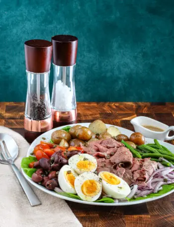 Roast beef Nicoise salad on a platter includes capers, eggs, deli sliced roast beef, tomatoes, olives, potatoes, onions, green beans, and a Dijon mustard dressing.