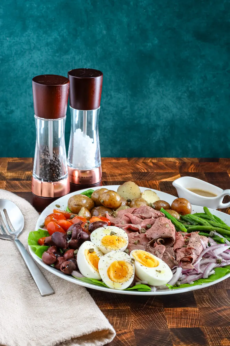 Roast beef Nicoise salad on a platter includes capers, eggs, deli sliced roast beef, tomatoes, olives, potatoes, onions, green beans, and a Dijon mustard dressing.