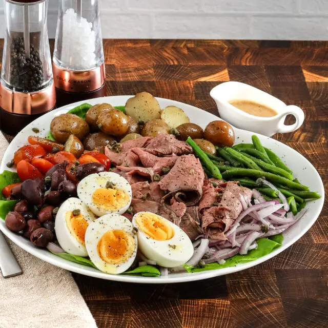 Roast beef nicoise salad arranged on a platter with eggs, deli sliced roast beef, tomatoes, olives, potatoes, onions, green beans, and a Dijon mustard dressing.