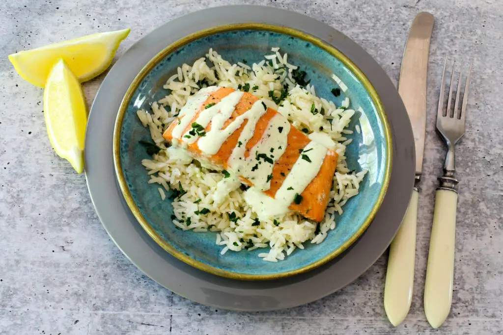 A plate with baked salmon on a bed of rice with a drizzle of basil mayonnaise and lemon wedges on the side.