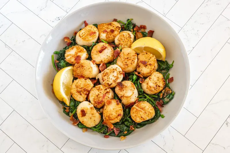 seared scallops in a serving dish on spinach with bacon and lemon wedges