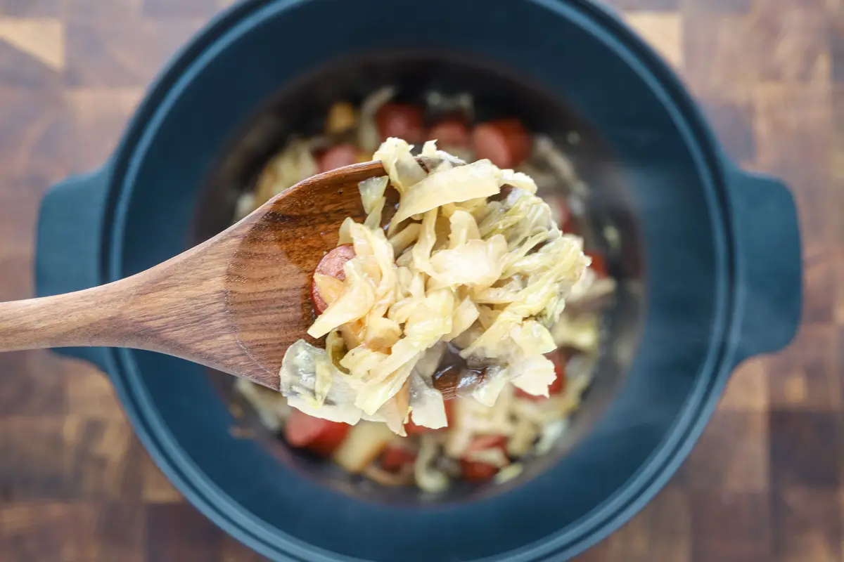 Slow cooker cooked kielbasa with apples, cabbage, onions, and tangy sauce, shown in the crock pot with a wooden spoon.