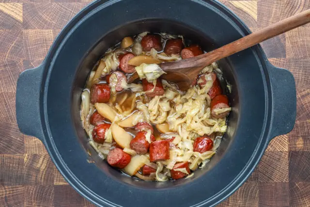 Slow cooker cooked kielbasa with apples, cabbage, onions, and tangy sauce, shown in the crock pot with a wooden spoon.