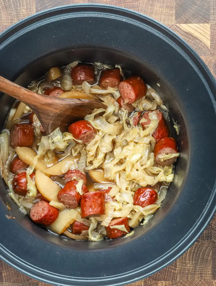 Slow cooker cooked kielbasa with apples, cabbage, onions, and tangy sauce, shown in the crock pot with a wooden spoon.