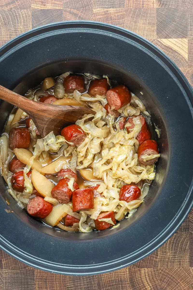 Slow cooker cooked kielbasa with apples, cabbage, onions, and tangy sauce, shown in the crock pot with a wooden spoon.