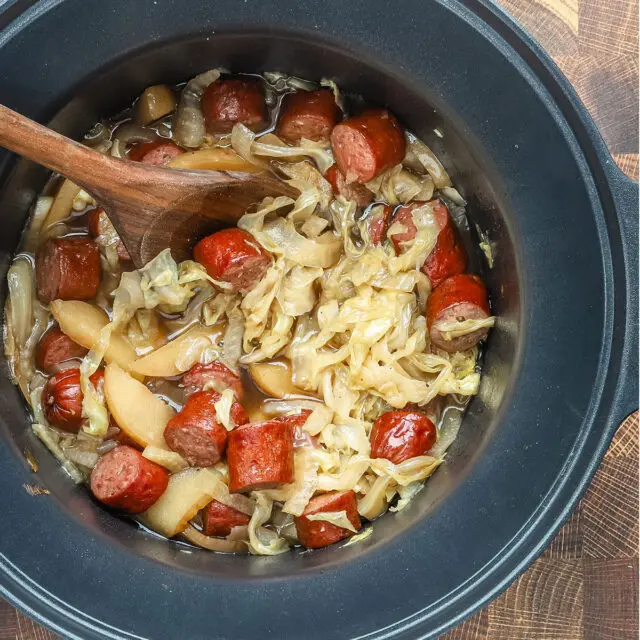 Slow cooker cooked kielbasa with apples, cabbage, onions, and tangy sauce, shown in the crock pot with a wooden spoon.