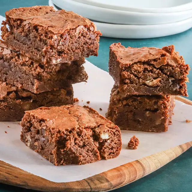 A stack of our ultimate fudgy brownies on a teak board with plates in the background.