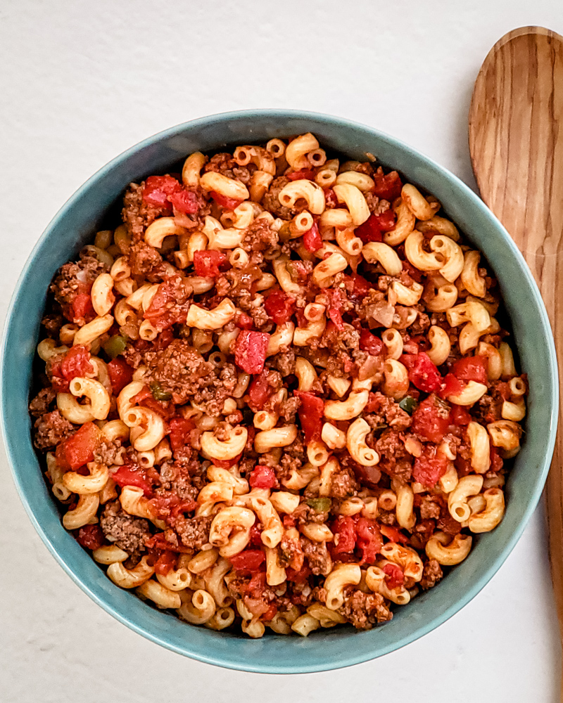 A serving bowl of American chop suey with a wooden spoon beside it.