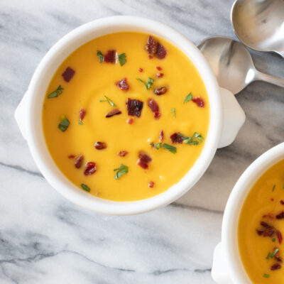 Bowls of butternut squash soup with bacon garnish on a marble slab with spoons beside them.