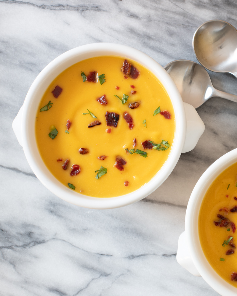 Bowls of butternut squash soup with bacon garnish on a marble slab with spoons beside them.