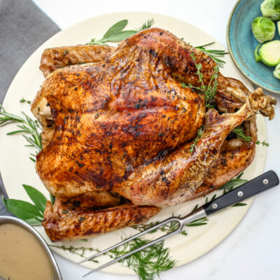 A whole roasted turkey with herbs surrounding it, and a dish of brussels sprouts on the side.