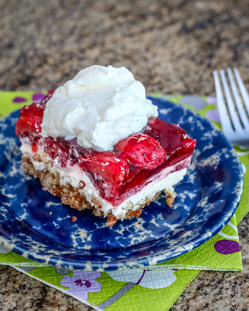 A strawberry dessert with a cream cheese layer and pretzel base, on a plate and topped with whipped topping.