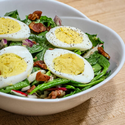 A bowl with spinach salad with bacon and sliced hard boiled eggs on top of it.
