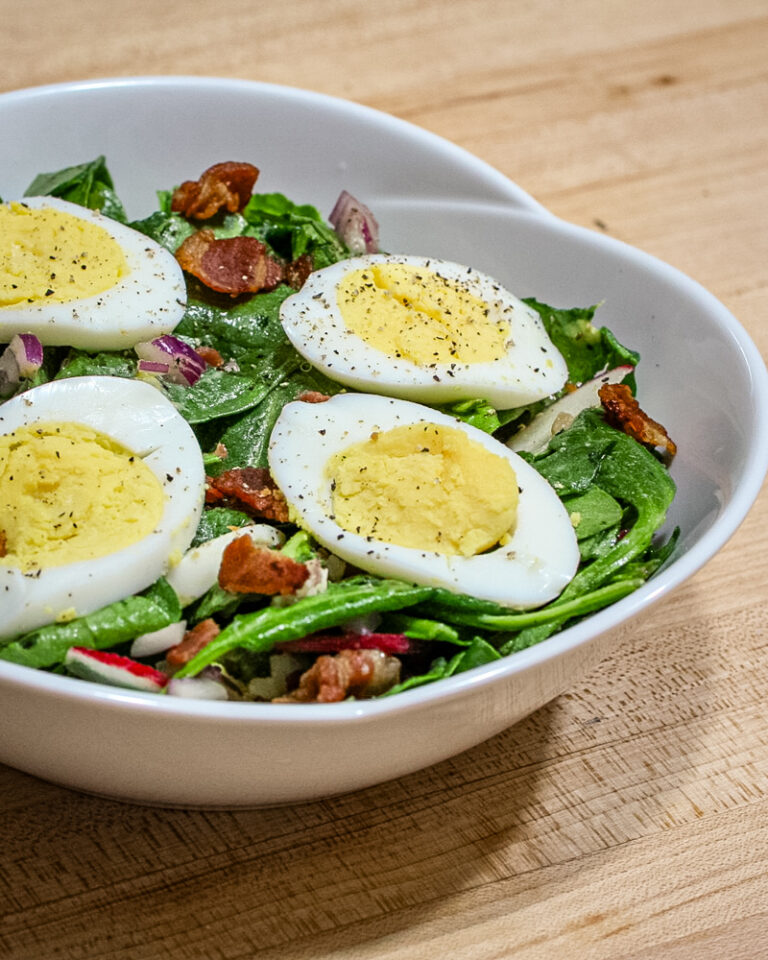 A bowl with spinach salad with bacon and sliced hard boiled eggs on top of it.