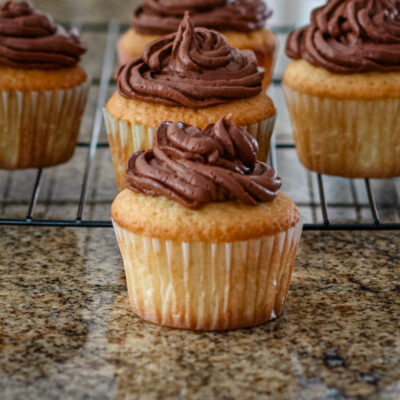 Moist vanilla cupcakes on a rack with chocolate frosting