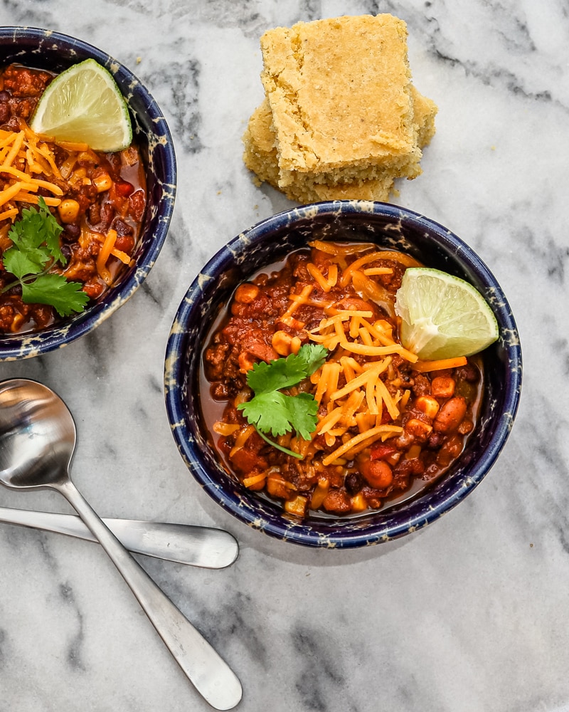 Beef and bean chili in bowls with garnishes and cornbread.