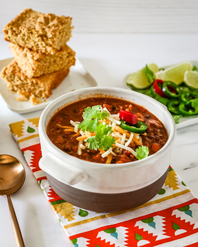 A bowl of turkey chili with garnishes and cornbread in the background.